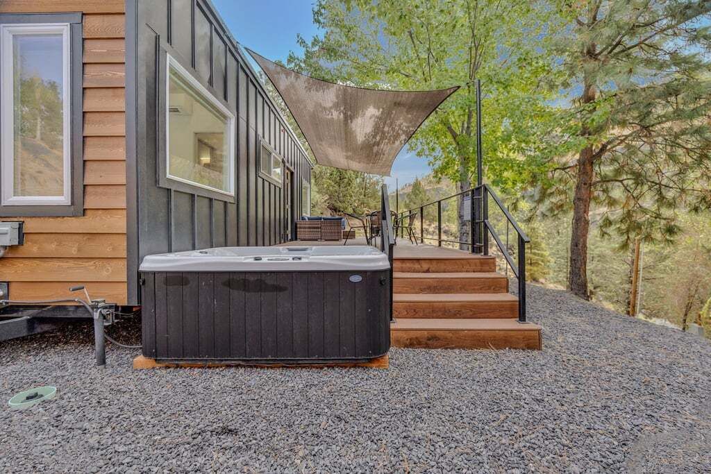 A hot tub outside a modern house with wooden siding, a large window, and a staircase leading up to a deck.