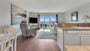 A modern, well-lit room with a flat-screen TV, a dining area, and a view of the sea through large windows.