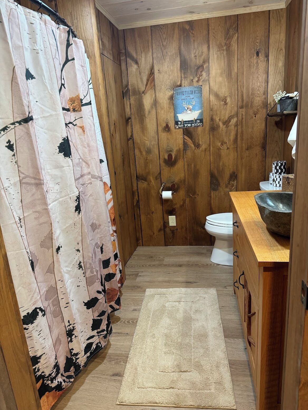 Un baño con paredes de madera, un inodoro, un lavabo con recipiente de piedra y una cortina de ducha con estampado floral.