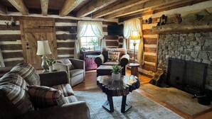 A rustic log cabin interior with a fireplace, wooden beams, a sofa, a coffee table, and a stone wall. - Log cabin with mountain VIEWS in Lexington! Near VA Horse Ctr, 8 mi from VMI/W&L (Lexington)