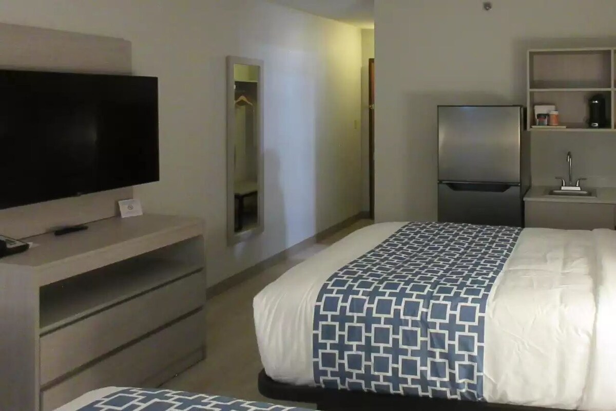 A hotel room with a bed, a TV on a stand, a refrigerator, and a small kitchenette area.