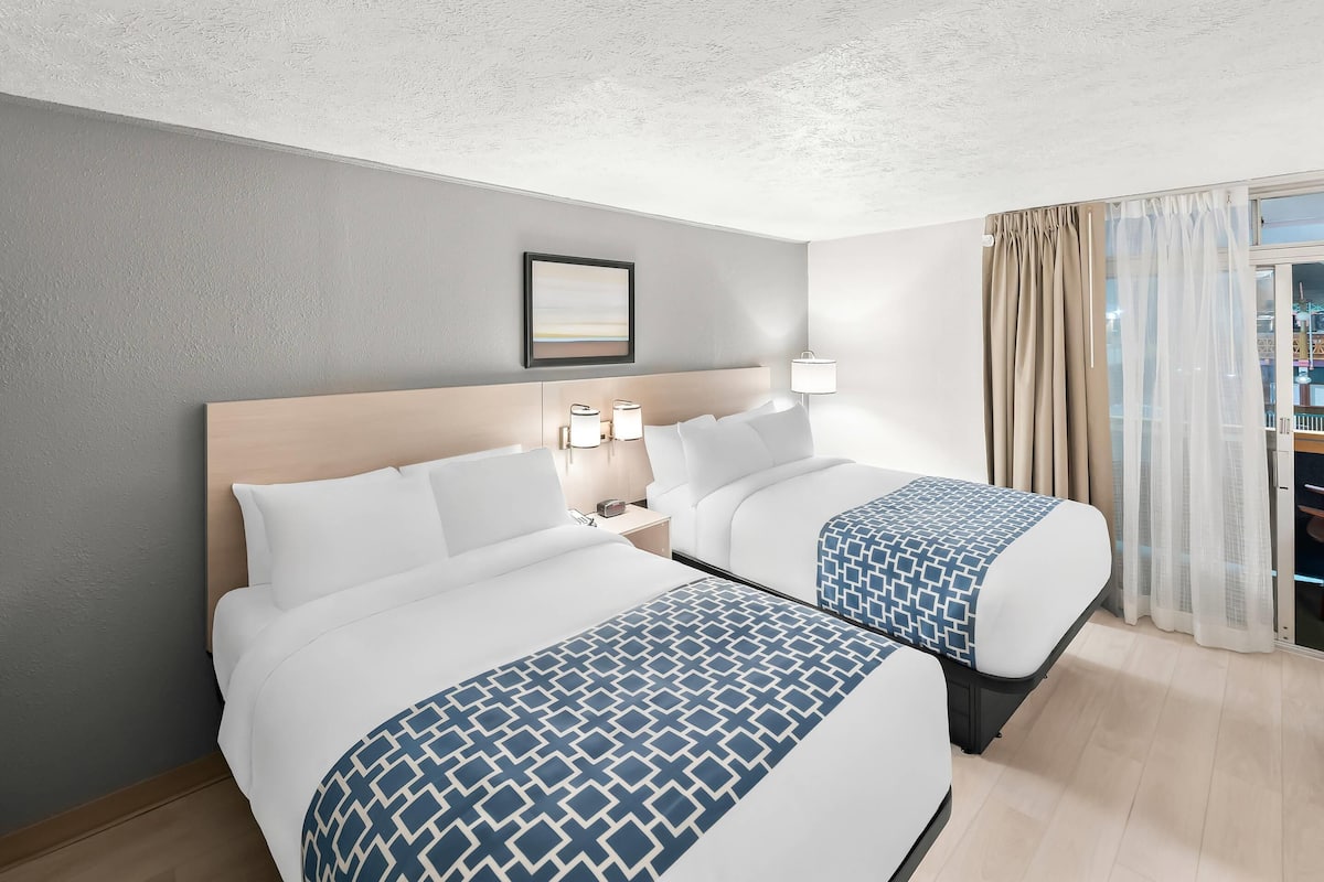 A hotel room with two beds, a bedside table with lamps, a framed picture on the wall, and a view through the window.