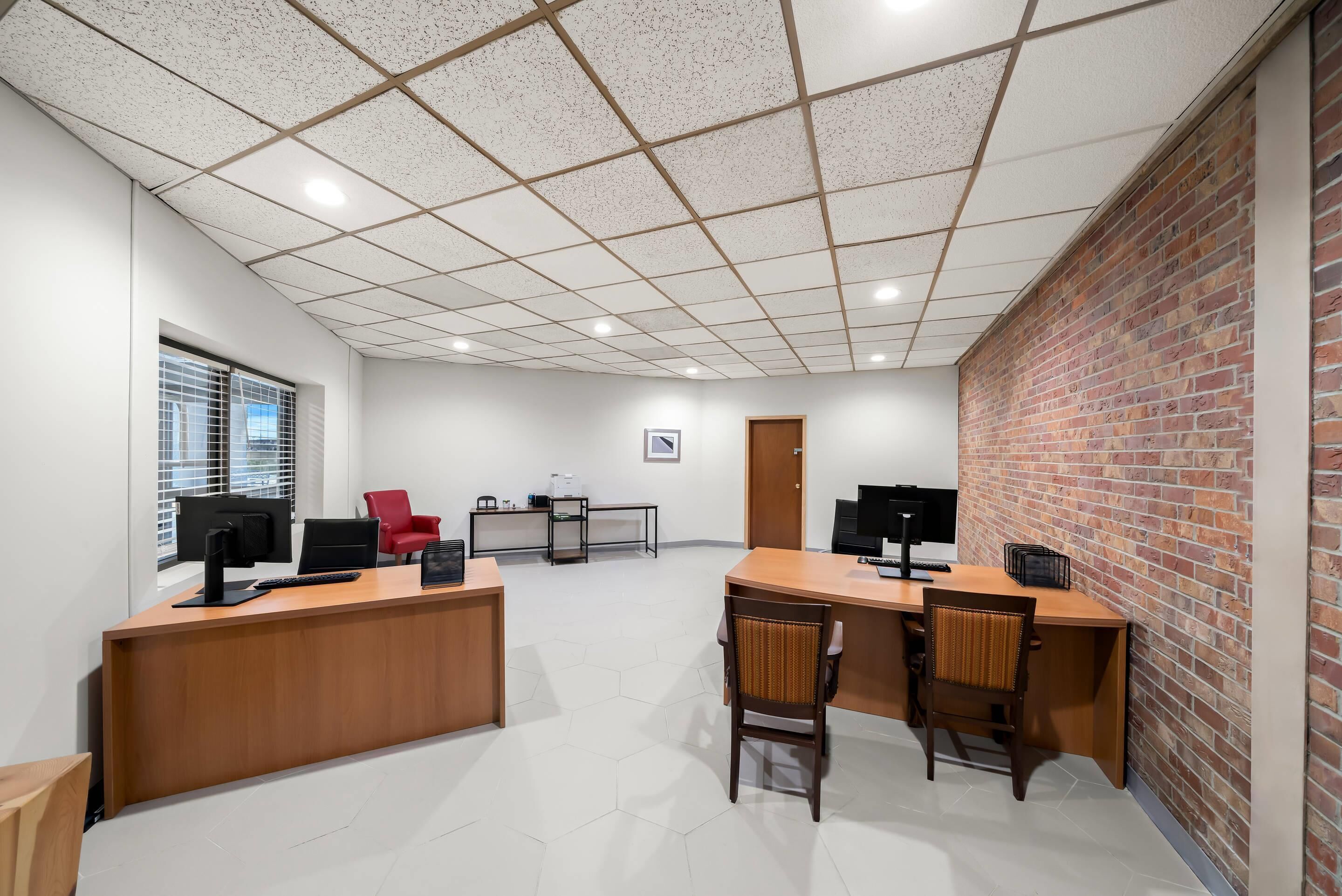 A spacious office with a wooden reception desk, two computer monitors, and a red chair. There is a brick wall on one side and a window with blinds on the other.