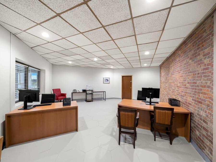 A spacious office with a wooden reception desk, two computer monitors, and a red chair. There is a brick wall on one side and a window with blinds on the other.