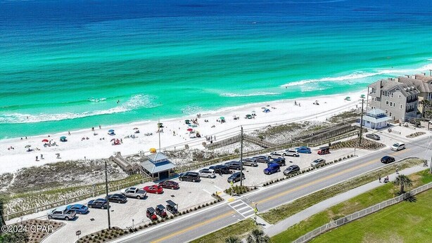 A beachside view with a parking lot, beach umbrellas, and a building with a balcony overlooking the ocean.