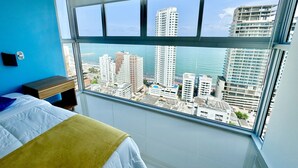A bedroom with a large window, a bed with a yellow blanket, a wooden bedside table, and a view of a cityscape and ocean. - Apartamento Navy 2 habitaciones (Cartagena)