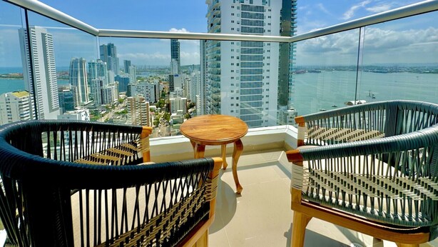 A balcony with wicker chairs and a small table, overlooking a cityscape with tall buildings and a body of water. - Apartamento Navy 2 habitaciones (Cartagena)