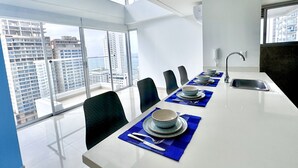 A modern dining area with a long table, blue placemats, and a city view. - Apartamento Navy 2 habitaciones (Cartagena)