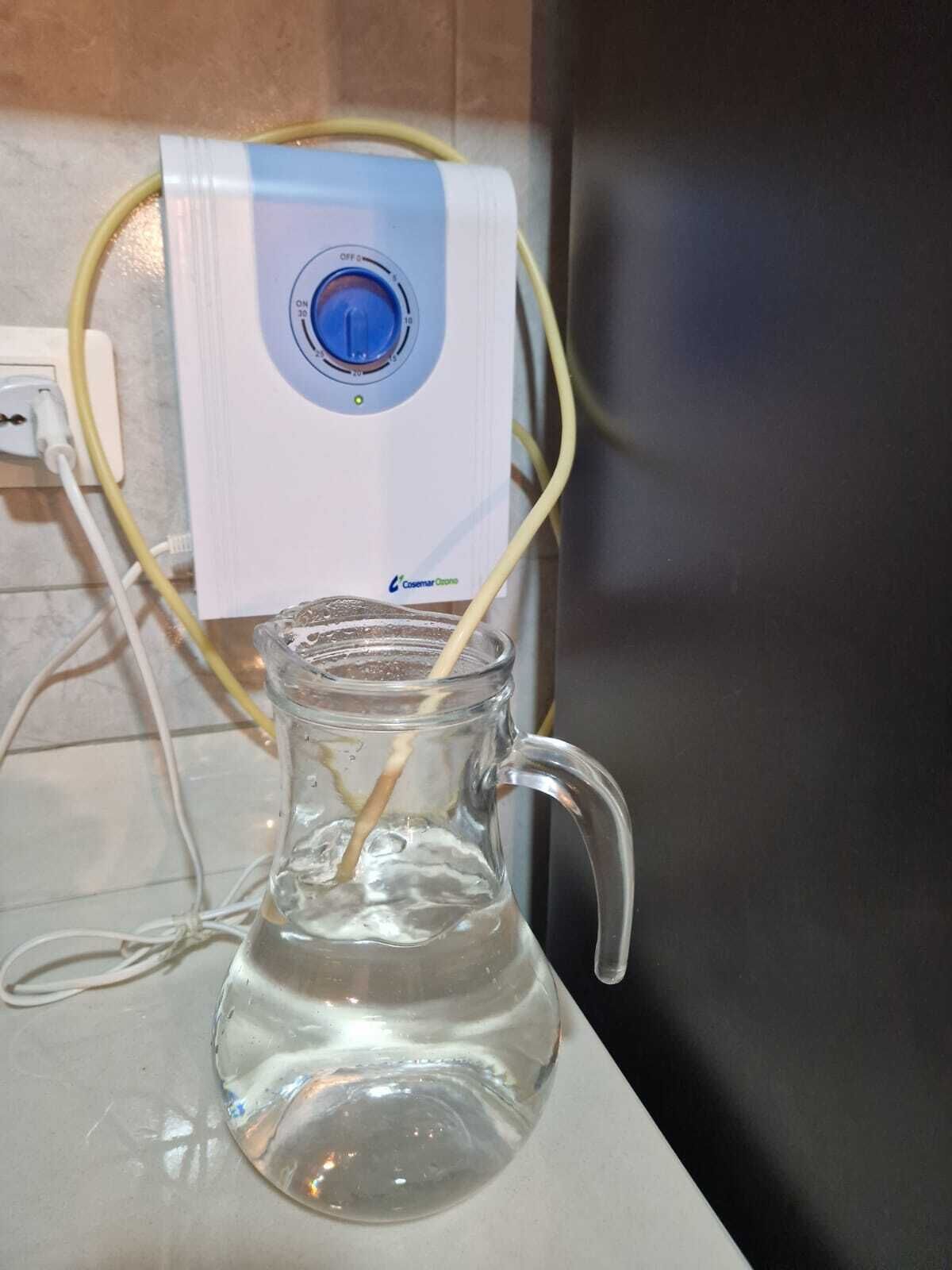 A clear glass pitcher with a handle, connected to a white device with a blue dial, possibly a water filter or purifier, on a countertop.