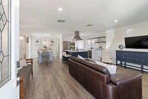 A modern kitchen with a central island, a dining area with a table and chairs, a brown leather sofa, and a TV mounted on the wall.