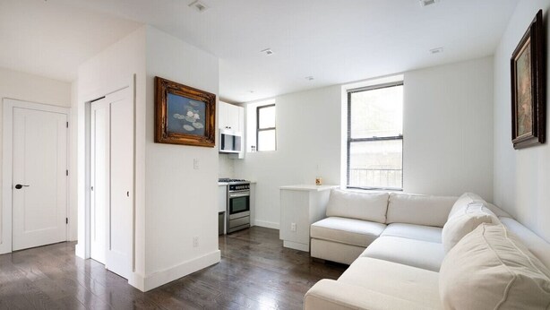 A spacious living room with a white sofa, a kitchen area with a stove and cabinets, and a large window providing natural light. - Prime soho 2 bedroom (New York)