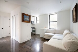 A spacious living room with a white sofa, a kitchen area with a stove and cabinets, and a large window providing natural light. - Prime soho 2 bedroom (New York)