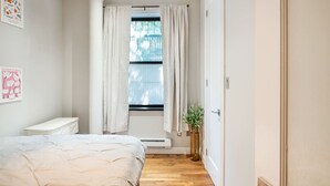 Une chambre à coucher avec un lit, une table de chevet, une fenêtre avec des rideaux, un radiateur et une porte.