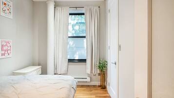 Un dormitorio con cama, mesita de noche, ventana con cortinas, radiador y puerta.