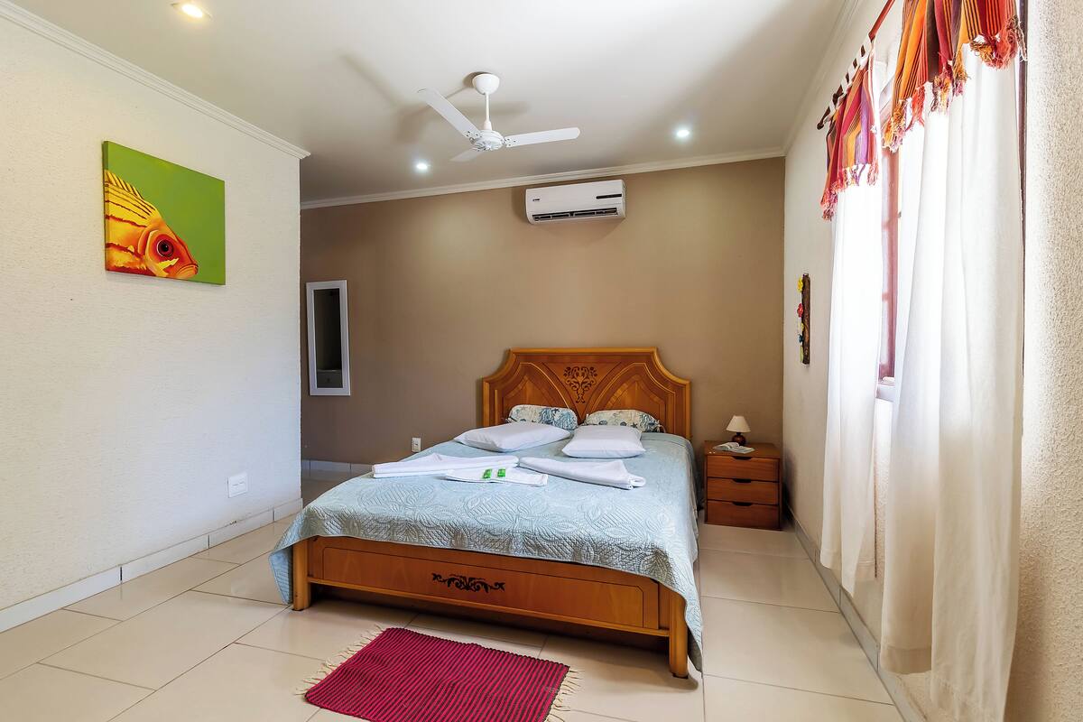 Una cama bien hecha con cabecera de madera, un ventilador de techo y un equipo de aire acondicionado en la pared.