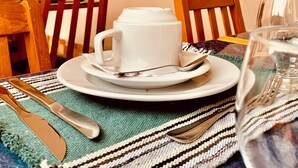 Une table à manger avec une tasse et une soucoupe blanches, un verre et des couverts. On y trouve deux chaises en bois, un chemin de table tissé et des décorations murales.