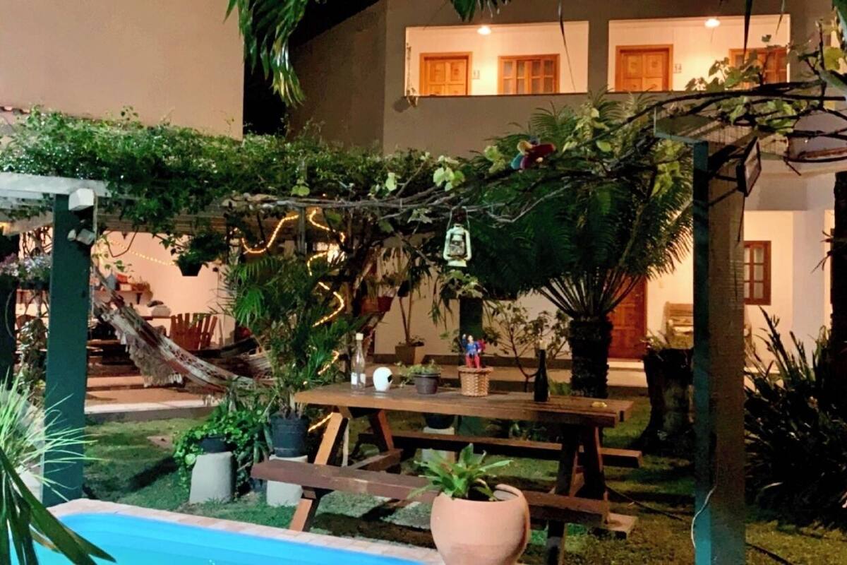 Un patio al aire libre con mesa y bancos de madera, rodeado de plantas en macetas y una pequeña piscina.