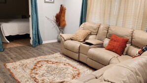 Un salon avec un canapé beige, un tapis à motifs et un vase contenant des plantes séchées.