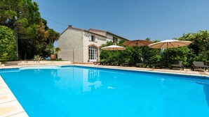 Una piscina con sillones de descanso y sombrillas, rodeada de follaje y un edificio con tejado de baldosas.