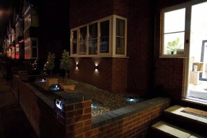 A nighttime view of a building with a window, a brick wall, and a small garden area with a water feature.