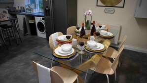 Zona pranzo moderna con tavolo in vetro apparecchiato per sei, circondato da sedie di legno. Sullo sfondo, si intravede un angolo cottura con lavatrice e un tavolo da pranzo con un vaso di fiori e bottiglie di vino.