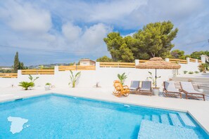 Une piscine entourée de chaises longues et dotée d’un parasol en chaume, le tout bordé de murs blancs et de verdure.