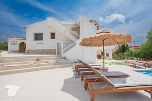 Une villa blanche à la façade en pierre, un escalier menant à l’entrée et un espace piscine agrémenté de chaises longues et d’un parasol en chaume.