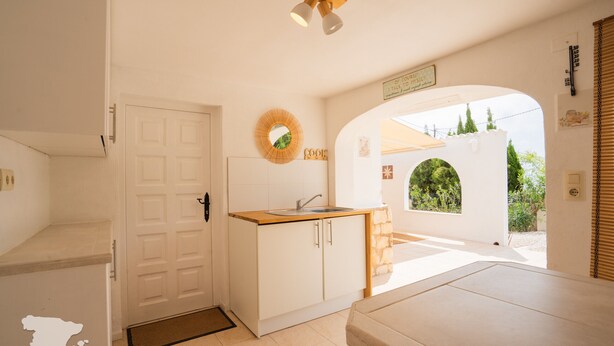 A compact kitchen with a sink, wooden countertop, and a small island. There is a door leading to another room and a view of greenery outside.