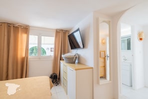 A bedroom with a bed, a TV mounted on the wall, a bedside table with a basket, a fan, and a view of a bathroom.