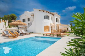 A white villa with a swimming pool, lounge chairs, and a dining area.