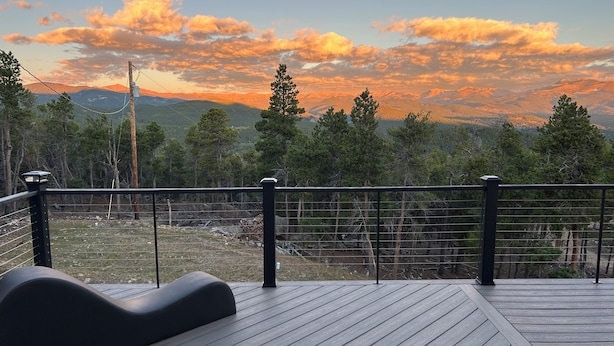 A wooden deck with a black railing, overlooking a mountainous landscape with trees and a sunset.