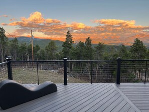 Ett trädäck med ett svart räcke, med utsikt över ett bergigt landskap med träd och en solnedgång.