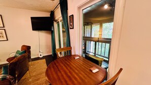A dining area with a wooden table and chairs, a television mounted on the wall, and a view of the outdoors through a window with curtains. - Forest Getaway & Golf at Lake (Bloomington)