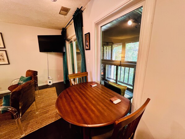 A dining area with a wooden table and chairs, a television mounted on the wall, and a view of the outdoors through a window with curtains.