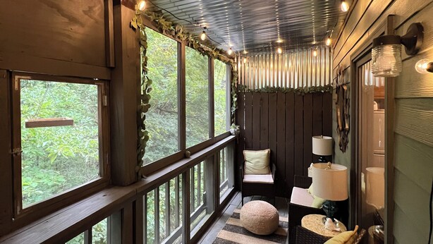 A covered porch with a wooden railing, a seating area with a cushioned chair and ottoman, and a view of greenery outside. - Forest Getaway & Golf at Lake (Bloomington)