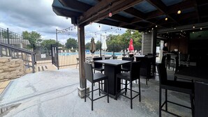An outdoor patio area with tables and chairs, a pool in the background, and a covered seating area. - Forest Getaway & Golf at Lake (Bloomington)