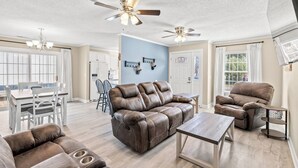 Un vaste salon avec un canapé en cuir marron, un fauteuil inclinable et une table basse en bois. On y trouve également des ventilateurs de plafond, un lustre et, au fond, un coin repas.