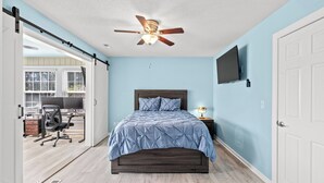 Une chambre à coucher comprenant un lit, un ventilateur de plafond, un téléviseur fixé au mur et un bureau avec une chaise dans la pièce adjacente.