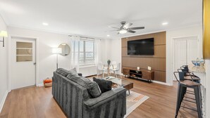 Un salon moderne avec un canapé gris, une table basse, un téléviseur fixé sur un mur en panneaux de bois, un ventilateur de plafond et un petit coin repas avec une table et des chaises.