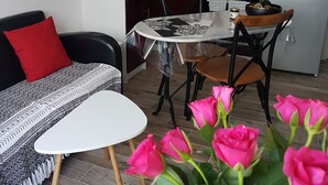 A modern kitchen with dark cabinets, a white countertop, and a stainless steel range hood. There is a dining area with a round table and chairs, and a sofa with a red pillow.