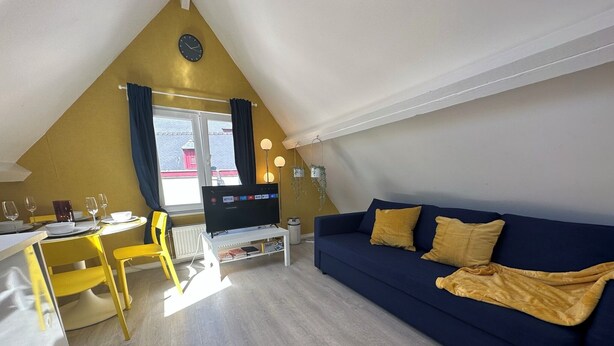 Una habitación acogedora con un sofá azul, sillas amarillas, una mesa de comedor y un televisor sobre un mueble blanco.