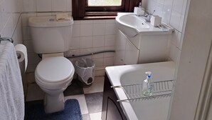 Une petite salle de bain avec des toilettes, un lavabo et une baignoire. Il y a une fenêtre avec du verre dépoli, une serviette accrochée au mur et une poubelle.