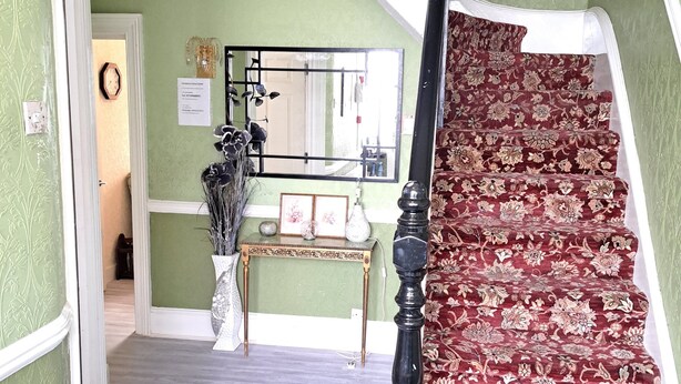 Un escalier recouvert d’une moquette à motifs rouges, avec des murs peints en blanc et une rampe en bois.