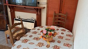 Un coin repas avec une table ronde recouverte d’une nappe à motifs floraux, entourée de chaises en bois. Un téléviseur est fixé au mur, au-dessus de la table.