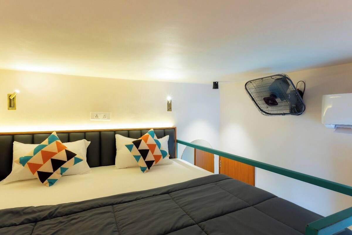 A hotel room with a bed, two pillows, a bedside table, a wall-mounted fan, and an air conditioning unit.