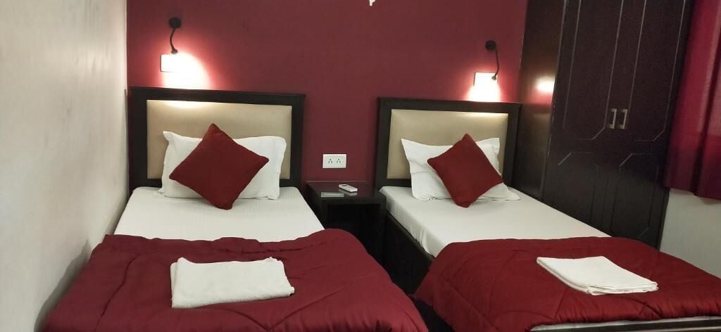 A hotel room with two single beds, each with a red bedspread and white pillows. There are bedside tables with lamps and a dark wooden wardrobe.