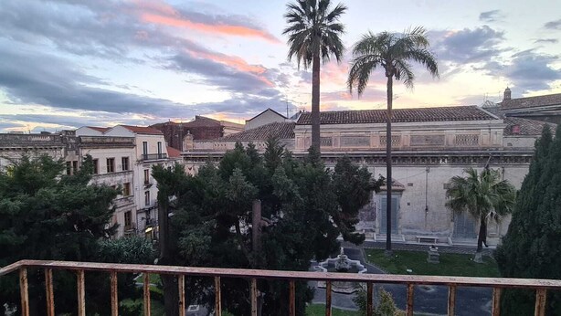 A courtyard with a fountain, palm trees, and historic buildings with tiled roofs. - Quiet and cozy in the center, one step away from everything and with a unique view (Catania)