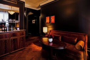 A bar area with a wooden bar counter, glasses, and bottles. There is a sofa with a vase of flowers and a lamp.