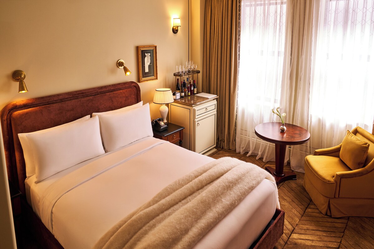 A hotel room with a large bed, a bedside table with a lamp, a small table with a vase, and a chair.