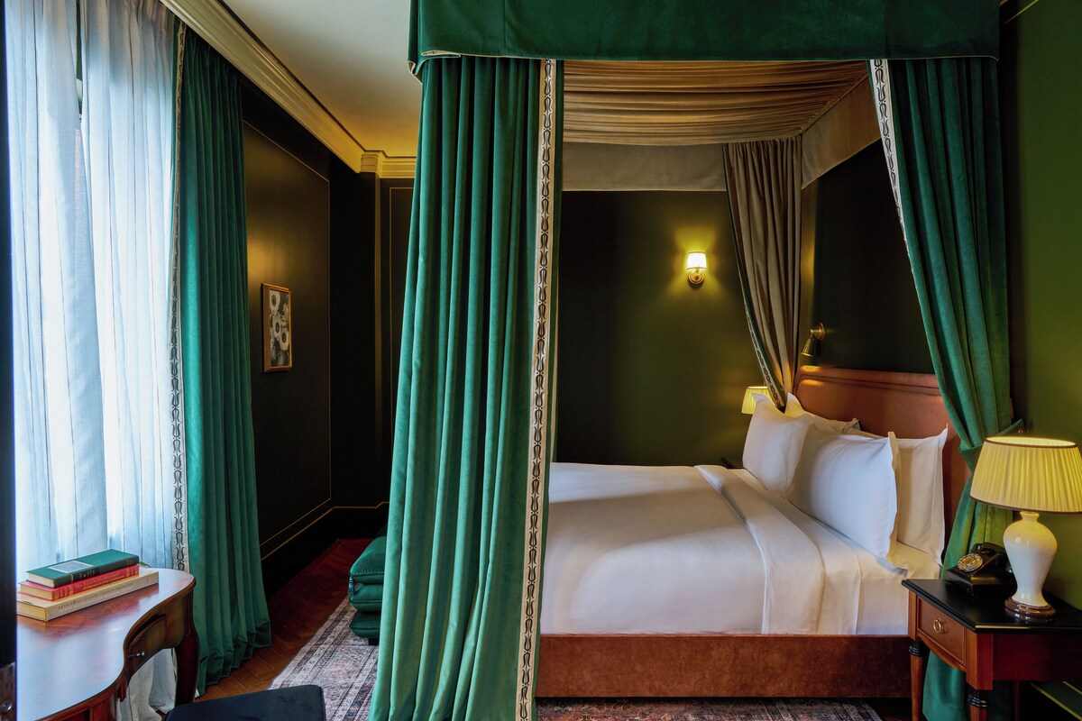 A four-poster bed with green curtains, a bedside table with a lamp, and a framed picture on the wall.